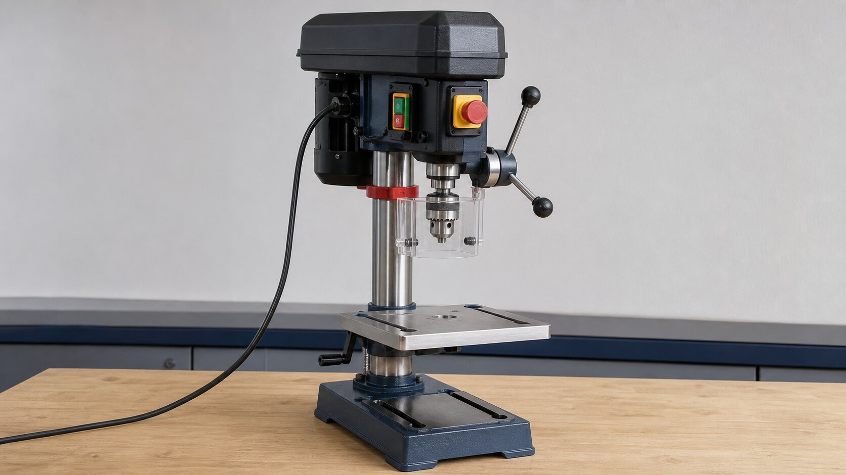 Bench-mounted drill press on a clean workshop bench with a guard, switches, power cable, table, and work area visible.