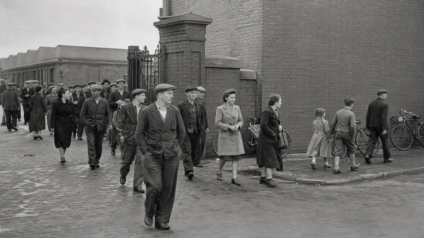AI-generated black-and-white historical reenactment of people leaving an industrial factory through a brick gate.