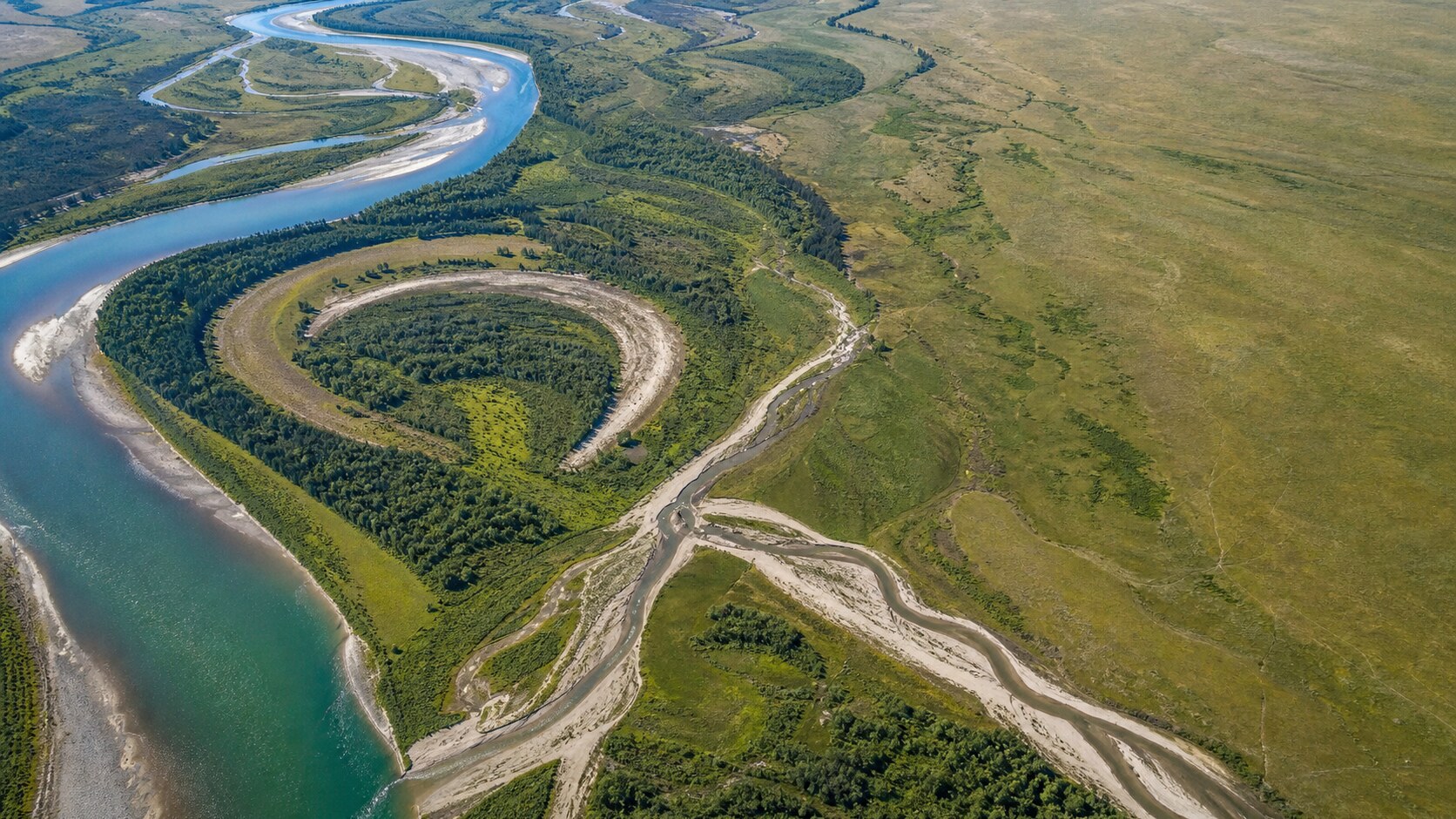 Aerial view of a winding river with broad meanders, exposed sediment bars, green floodplain, and eroded banks.
